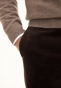 Bruin textuurtrui met geribbelde manchetten, gecombineerd met donkere corduroybroeken met een zijzak. Schone witte shirtmanchet zichtbaar eronder.