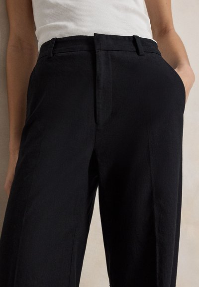 Άτομο που φοράει μαύρο παντελόνι ψηλόμεσο και λευκό αμάνικο τοπ, με το ένα χέρι στην τσέπη, πάνω σε ουδέτερο φόντο.