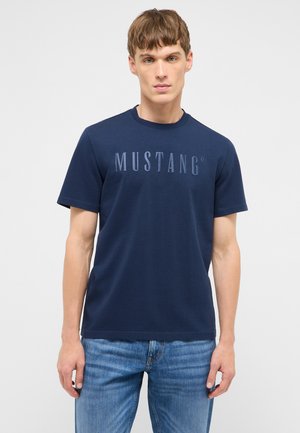 Junger Mann, der ein dunkelblaues T-Shirt mit Mustang-Logo und hellblaue Jeans trägt, steht vor einem einfarbigen hellgrauen Hintergrund.
