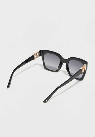 Μαύρα γυαλιά ηλίου με τετράγωνο πλαίσιο, βαθμιαία σκουρόχρωμους φακούς και χρυσά διακοσμητικά μεντεσέδες που ακουμπούν σε λευκή επιφάνεια.