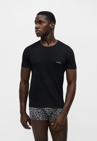 Άνδρας που φοράει μαύρο t-shirt Hugo και σορτς με ασπρόμαυρο σχέδιο, κοιτάζει προς τα αριστερά σε απλό φόντο.