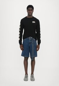 Svart langærmet skjorte med logo, denimshorts med øjenlidsdetaljer og grå sneakers. Model står mod en hvid baggrund.