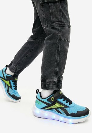 Dítě oblečené v černých kalhotách typu cargo a modrých teniskách Reebok se svítící podrážkou na bílém pozadí.