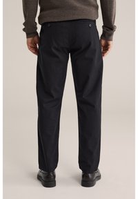 Hombre vistiendo pantalones negros de pierna recta, suéter marrón oscuro y zapatos negros, de pie de espaldas contra un fondo liso.