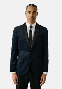 Jachetă de tuxedo albastru marin cu model, rever din satin, închidere cu un singur nasture și două buzunare, purtată peste o cămașă albă și o cravată neagră.