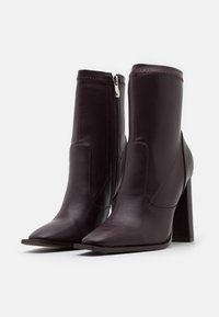 Par de botas de cuero de color púrpura oscuro hasta el tobillo con cremallera lateral, puntera afilada y tacón alto en bloque sobre un fondo blanco.