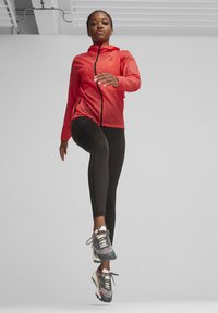 Piros, könnyű dzseki kapucnival és fekete leggings; mintás, párnázott sportcipők fűzős kivitelben; a modell dinamikus pózban van.
