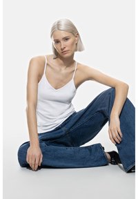 Top bianco a spalline abbinato a jeans denim blu larghi. Il modello è seduto a gambe incrociate su uno sfondo chiaro. Design semplice, senza pattern.