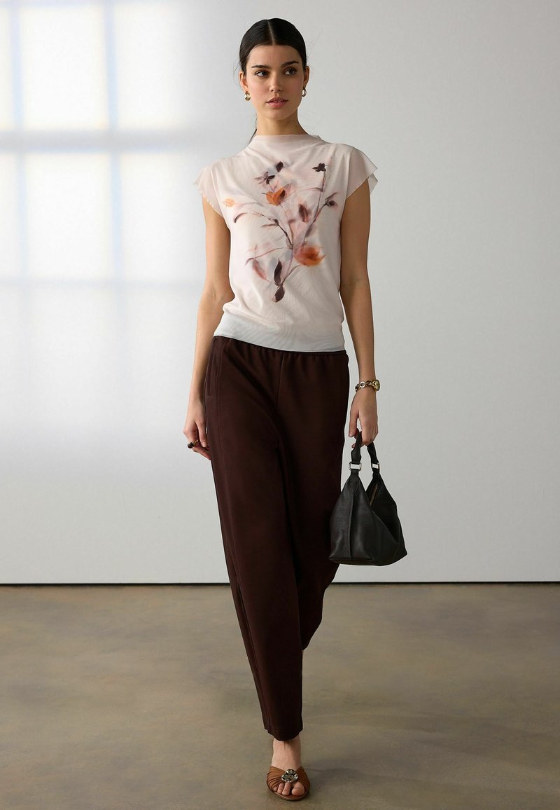 Jeune femme aux cheveux tirés en arrière portant un haut fleuri pastel, un pantalon sombre et ample, des sandales marron, avec un sac à main noir à l'intérieur.