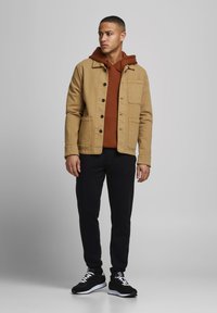 Giacca beige con bottoni neri sopra un hoodie marrone, abbinata a pantaloni neri e sneakers nere. Design semplice, tessuto di peso medio.