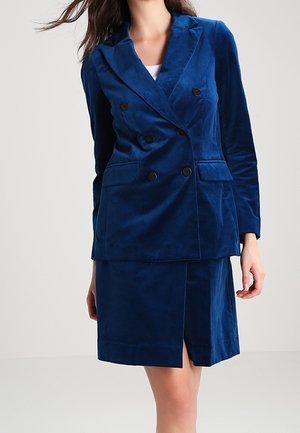 Femme portant un blazer en velours bleu cintré à double boutonnage avec des boutons noirs et une jupe assortie longueur genou avec une fente à l'avant.