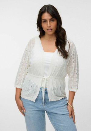 Donna con capelli lunghi e scuri che indossa un cardigan bianco a maglia aperta legato in vita sopra una maglietta bianca e jeans azzurri, in piedi su uno sfondo semplice.