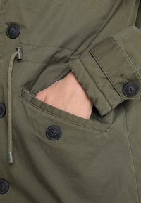 Olivgrön jacka i texturerat tyg, med svarta knappar, dragsko i midjan och sidofickor.