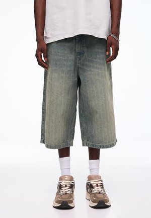 Osoba nosi vybledlé džínsy s širokými nohavicami, biele ponožky s krátkym lemom, béžovo-hnedé tenisky a bielu textúrovanú košeľu s krátkymi rukávmi.