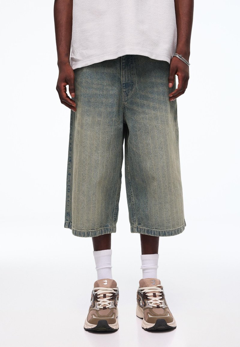 Personne portant un short en jean large délavé, des chaussettes blanches montantes, des baskets beige et marron, et une chemise blanche texturée à manches courtes.