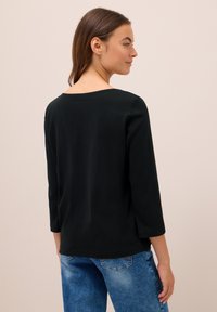 Frau mit braunen Haaren, die ein schwarzes, geripptes Langarmoberteil und blaue Jeans trägt, seitlich vor einem schlichten beigen Hintergrund steht.