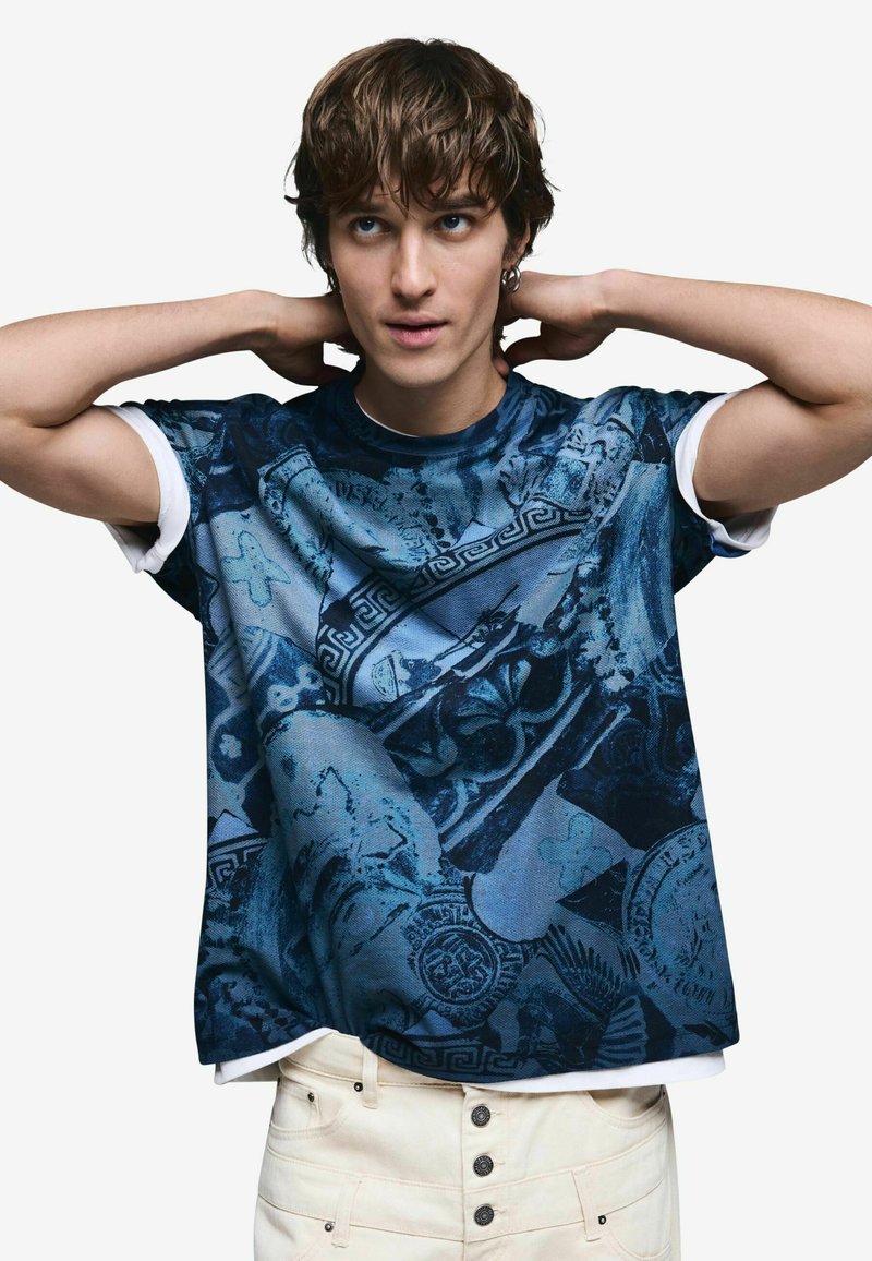 Modré grafické tričko s krátkymi rukávmi, zdobené zložitými vzormi a dizajnmi, s pevným bielym okrúhlym výstrihom a textúrovanou tkaninou.