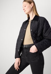 Jachetă neagră matlasată cu nasturi tip snap și guler din catifea. Pur tată deasupra unei bluze bej și a unor blugi negri, evidențiind buzunarele și designul elegant.