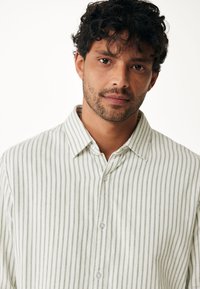 Světle zelená a bílá pruhovaná košile na knoflíky s límečkem a jemnou texturou. Přední knoflíky jsou viditelné, s uvolněným střihem.