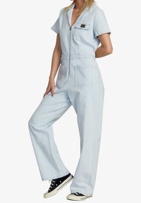 Jumpsuit rayé bleu clair avec des manches courtes, fermeture éclair sur le devant, taille cintrée et deux poches sur la poitrine. Accompagné de baskets noires.