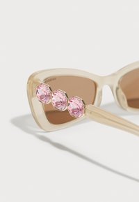 Óculos de sol estilo gato com uma armação transparente em bege, lentes castanhas e três realces em cristal rosa na parte da frente.