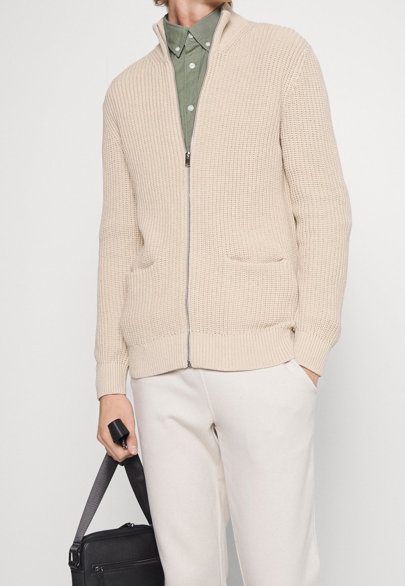 Béžový svetr na zip s texturovaným pletením, dvěma předními kapsami, ribovanými manžetami a lemem. Nosený přes zelenou košili na knoflíky, držený s černou taškou.