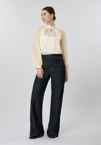 Kvinde i beige langærmet bluse med nøglehulshalsudskæring og sort, højtsiddende bukser med vide ben, stående og kiggende mod venstre.