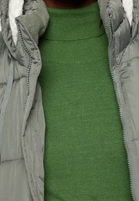 Chaleco acolchado gris con un suave forro interior ligero sobre un jersey verde con cuello alto acanalado. Detalles en primer plano muestran la textura y la cremallera del chaleco.