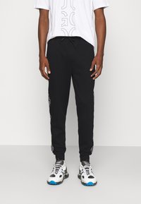 Osoba oblečená v černých teplácích s bočními grafickými detaily, bílém tričku s grafikou a bílých, černých a modrých teniskách stojící před světlým pozadím.