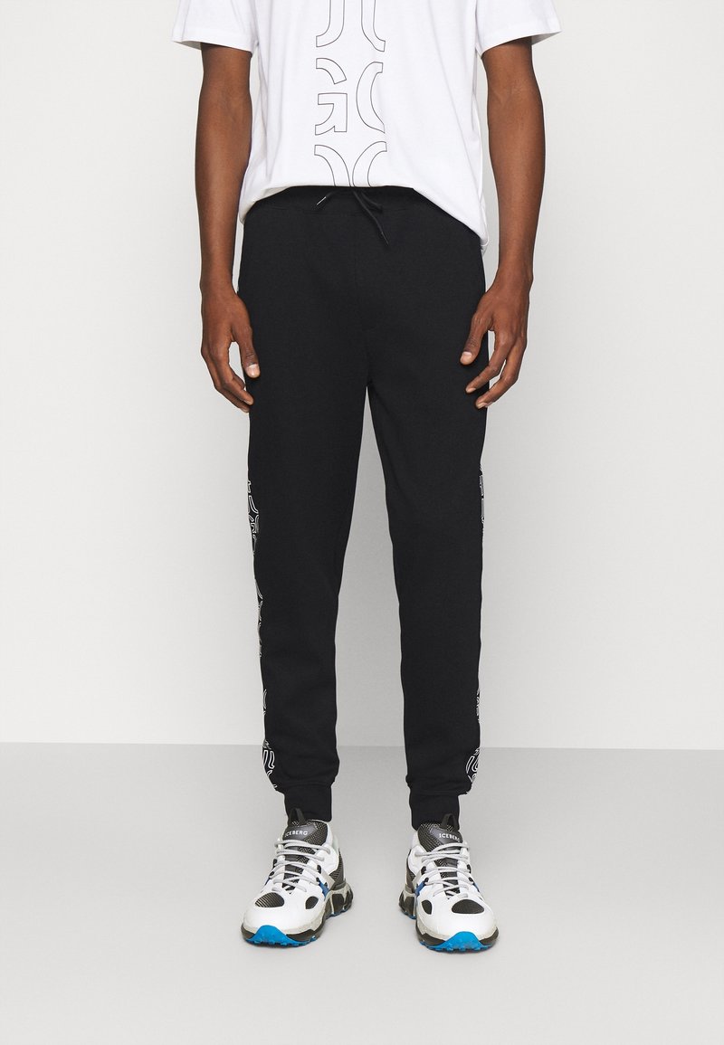 Osoba oblečená v černých teplácích s bočními grafickými detaily, bílém tričku s grafikou a bílých, černých a modrých teniskách stojící před světlým pozadím.