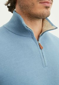 Veste zippée bleu clair avec un col haut, dotée d'une doublure beige et d'un tissu texturé. Comprend un accent en cuir sur la tirette du zip.