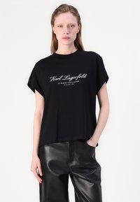 Juodas medvilninis marškinėlis trumpomis rankovėmis, su baltu siuvinėtu logo „Karl Lagerfeld“ priekyje. Laisvas siluetas ir lygus paviršius.