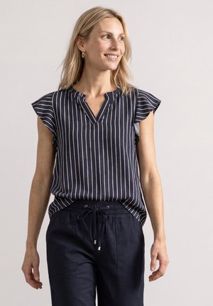 Femme blonde portant un chemisier bleu marine à rayures verticales et un pantalon bleu marine à cordon, souriante et regardant vers la droite sur un fond uni.