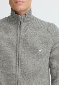 Pelēks ribots adījums džemperis ar augstu apkakli un pilnu rāvējslēdzēju. Iezīmē neliels balts simbols uz krūtīm un teksturēta auduma.
