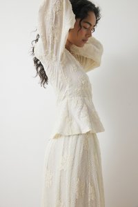 Blusa bianca in pizzo con maniche a sbuffo e ricami floreali, abbinata a una gonna in pizzo coordinata. Tessuto testurizzato con dettagli di design a strati.