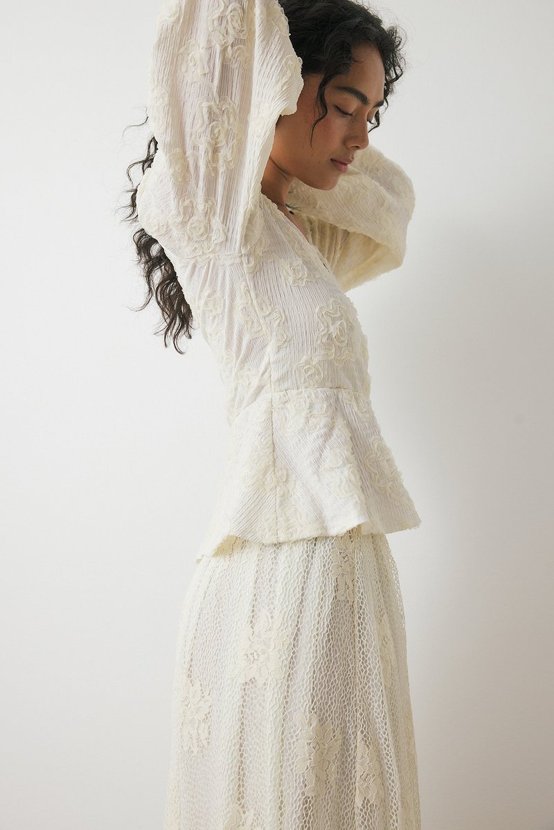 Blusa bianca in pizzo con maniche a sbuffo e ricami floreali, abbinata a una gonna in pizzo coordinata. Tessuto testurizzato con dettagli di design a strati.