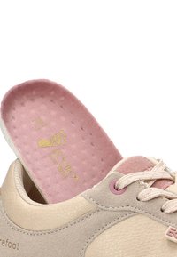 Zapatilla de color beige claro con cordones crema y una plantilla acolchada rosa parcialmente retirada, mostrando los detalles de tamaño y logo en el interior del zapato.