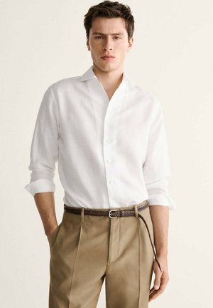 Joven con camisa blanca de manga larga remangada metida en pantalones beige con un cinturón trenzado marrón, de pie con las manos en los bolsillos.