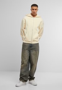 Lyscremet zip-up hoodie med en afslappet pasform, der har en stor hætte og kengurulomme, parret med løse, slidte grå jeans og hvide sneakers.