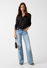 Schwarzes, langärmliges Hemd mit Knopfleiste, kombiniert mit hellblauen, weiten Jeans. Die Accessoires umfassen eine schwarze Handtasche und einen silbernen Gürtel.