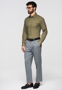 Homme avec barbe et lunettes portant une chemise boutonnée vert olive, un pantalon gris plissé, une ceinture noire et des mocassins noirs, debout devant un fond uni.