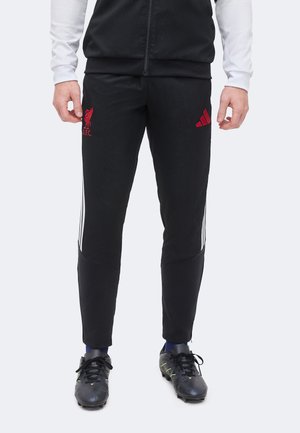Bărbat purtând pantaloni de antrenament negri Liverpool FC cu logo-uri roșii, ghete de fotbal negre, șosete bleumarin și un pulover alb cu mâneci lungi pe fundal alb.