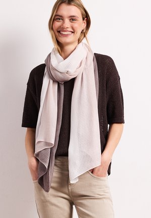 Femme souriante portant un haut en maille foncé, un pantalon beige et une grande écharpe dégradée claire et foncée gris drapée autour du cou.