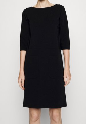 Zwarte knielange jurk met driekwartmouwen en twee opgestikte zakken aan de voorkant, gedragen door een staande persoon, neutrale achtergrond.