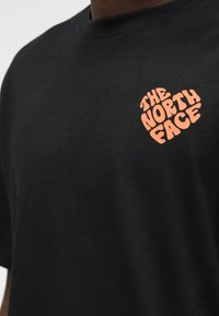 Μαύρο μπλουζάκι με ένα μικρό πορτοκαλί λογότυπο σε σχήμα καρδιάς που γράφει "THE NORTH FACE" στην αριστερή πλευρά του στήθους.