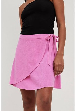 Kobieta ubrana w różową spódnicę zawiązaną w pasie oraz czarny top bez rękawów, stojąca na tle jasnego, jednolitego tła.