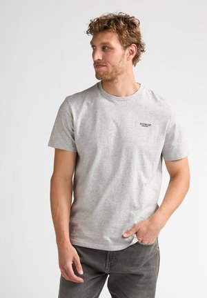 Man met krullend haar die een lichtgrijs T-shirt met korte mouwen met spikkels draagt en donkergrijze broek, kijkt zijwaarts met één hand in de zak.