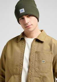Olijfgroene gebreide beanie en tan werkjack met een patchzak, voorzien van een vierkant logo. Wit t-shirt steekt uit de jas.