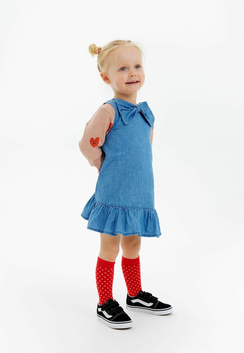 Denim mekko, jossa on nauhatakki ja rimsupohja, yhdistettynä pitkähihalaiseen vaaleanpunaiseen paitaan, jossa on sydänkuvioita, sekä punavalkoisiin pilkullisiin polvisukkiin.