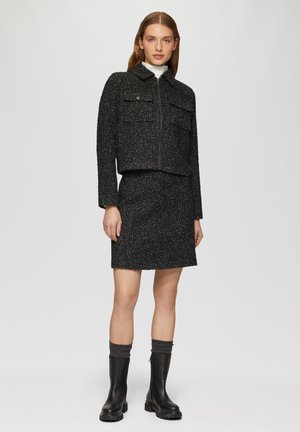 Zwarte tweed cropped jas met twee voorzakken, gecombineerd met een bijpassende rok. Model draagt kniehoog zwarte laarzen met grijze manchetten.